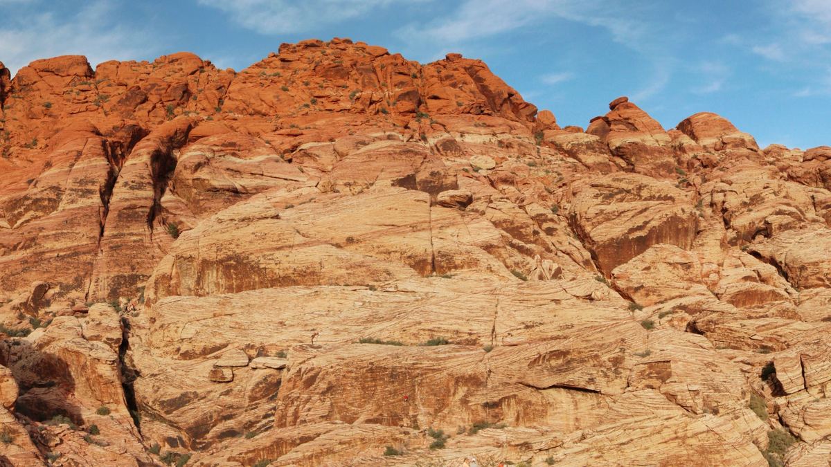 Wide panorama of a rugged red and orange rock hill with desert scrub in the foreground under a blue sky with thin clouds.